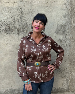 A woman wearing a brown long-sleeve button-up shirt with a white horse print, styled with a wide brown belt featuring a large turquoise and gold concho buckle, paired with blue jeans, standing against a textured concrete wall.