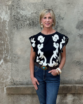 Blonde woman against a concrete wall in a western black and creme  print top and jeans