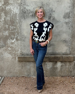 Blonde woman against a concrete wall in a wester black and creme  print top and jeans