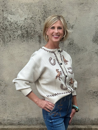 Western inspired cream cardigan with knit horses, horseshoes, zigzag pattern, and brown whipstitch trim on neckline and button placket