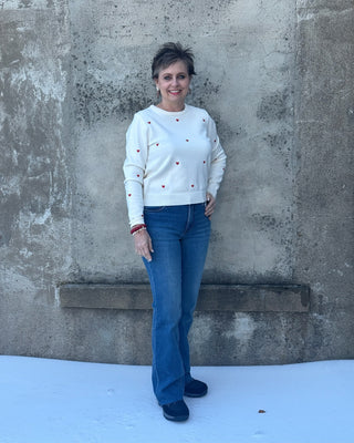 Woman wearing a white sweater with red accents and blue jeans standing against a textured wall.