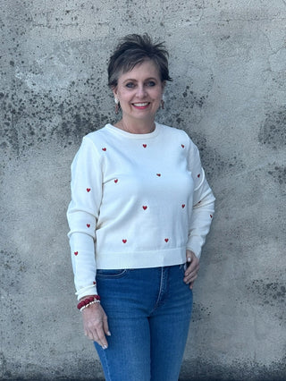 Woman wearing a white sweater with red accents and blue jeans standing against a textured wall.