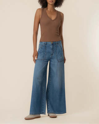 A model wearing a brown sleeveless V-neck top and high-waisted, wide-leg blue denim jeans, standing against a plain beige studio background.