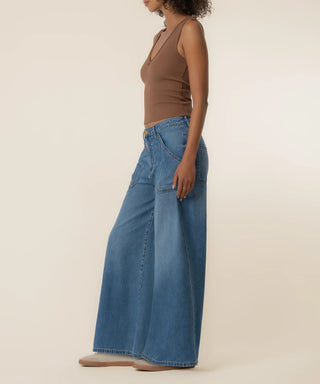 A model shown from the side wearing a pair of wide-leg blue denim jeans and a brown sleeveless top, standing against a plain beige studio background.