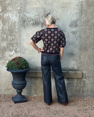 Person wearing a patterned top and dark pants standing against a textured wall with a planter nearby.