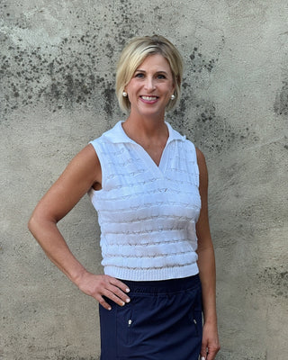 Woman wearing a light blue sleeveless top and dark blue pants against a textured wall.