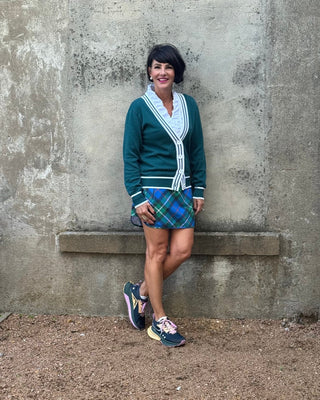 Woman in a green cardigan and plaid skirt leaning against a concrete wall.