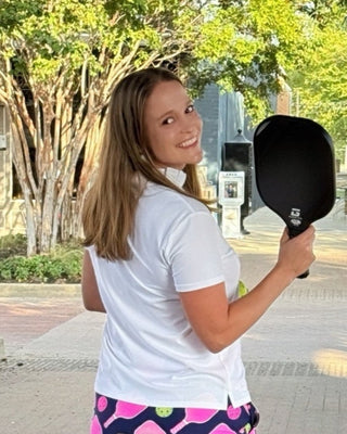 Person holding a pickleball paddle on a sidewalk with trees and buildings in the background