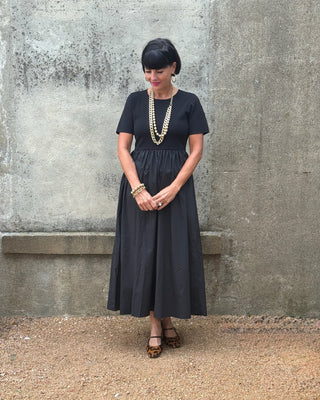 Woman wearing a black dress with layered necklaces against a gray wall.