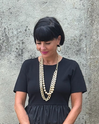Woman wearing a black dress with layered necklaces against a gray wall.