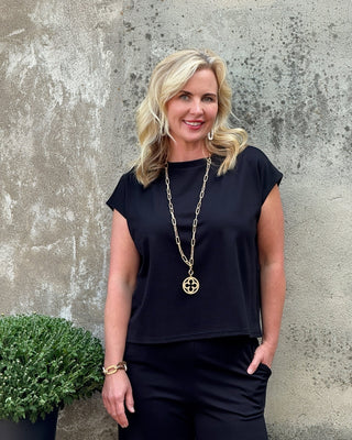 Woman wearing a black top and gold necklace against a textured wall.