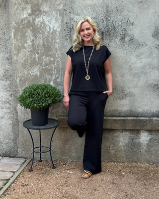 Woman in black outfit standing next to a potted plant against a textured wall.